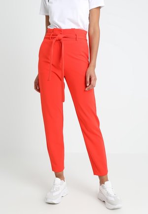 Mujer vistiendo pantalones de talle alto de color rojo brillante con un cinturón atado, combinados con una camiseta blanca y zapatillas blancas sobre un fondo liso.