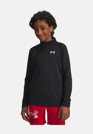 Mlada oseba, oblečena v črn športni zgornji del z zadrgo do četrtine in rdeče kratke hlače z logotipi Under Armour, se smeji in stoji na preprostem ozadju.