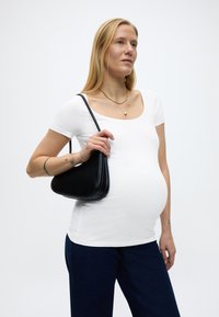 Trudna žena u bijeloj uskoj majici i tamnim trapericama, drži crnu torbu preko ramena, stoji ispred jednobojne svijetle pozadine.