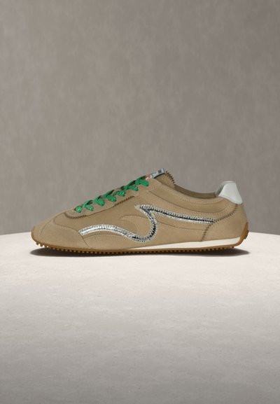 Μπεζ σουέντ αθλητικό παπούτσι με πράσινα κορδόνια, ασημί κυματιστό σχέδιο στο πλάι, λευκή ταμπέλα στη φτέρνα και καφέ σόλα πάνω σε ανοιχτή επιφάνεια.
