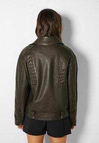 Brun konstläderjacka med klassisk krage, vertikala sömmar och dragkedjor vid ärmsluten. Slät yta, figursydd design och ingen synlig hårdvara.