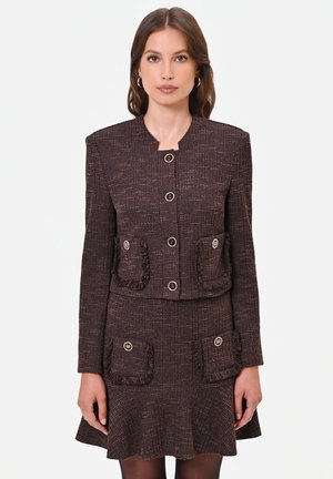 Donna che indossa un completo marrone scuro strutturato con giacca abbottonata e gonna abbinata con tasche frontali e dettagli con frange.