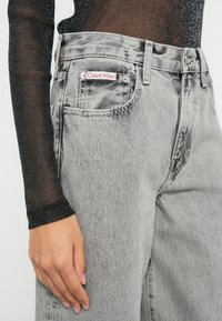 Šedé rifle Calvin Klein s vysokým pásom a vybledlou textúrou, s červenou logom značky na zadnom vrecku a detailmi gombíkov na páse.