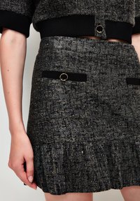 Zwarte textuurskirt met metallic draden, voorzien van een gerimpelde zoom, voorzakken met zwarte accenten en details in goudkleurig beslag.