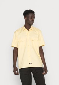 Camisa de manga curta, com botões, em amarelo claro, apresentando dois bolsos frontais, um colarinho clássico e uma etiqueta de logo preta na bainha.
