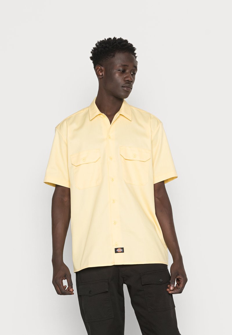 Camisa de manga curta, com botões, em amarelo claro, apresentando dois bolsos frontais, um colarinho clássico e uma etiqueta de logo preta na bainha.
