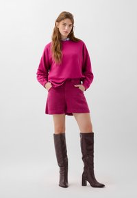 Fuchsia sweatshirt met lange mouwen, gelaagd over bijpassende fuchsia short. Kniehoge bruine leren laarzen met vierkante hakken en een gladde textuur.