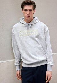 Gray hoodie with "BOULEVARD HAUSSMANN" in light yellow text, featuring a front pocket and drawstring hood, paired with navy pants.