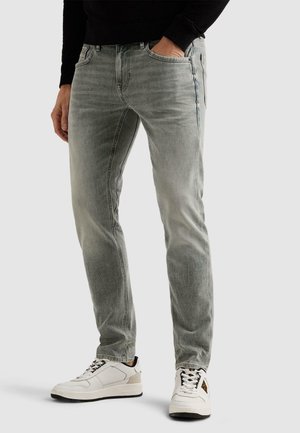 Moški, oblečen v izprane sive kavbojke, bele superge in črno majico z dolgimi rokavi, z eno roko v žepu, stoji na enobarvnem ozadju.