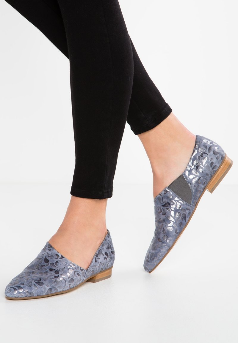 Pieds chaussés de mocassins à bouts pointus, à motifs argentés et bleus, avec de petits talons en bois, assortis à un pantalon noir ajusté.