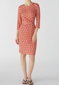 Robe moulante jusqu'aux genoux en rouge avec un motif géométrique blanc, dotée d'un décolleté rond et de manches trois-quarts avec un détail de taille nouée.