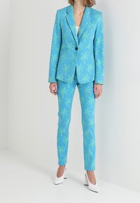 Kobieta ubrana w dopasowany niebieski garnitur z zielonymi wzorami palm, białą bluzkę pod spodem i białe buty na wysokim obcasie, stojąca na białym tle.