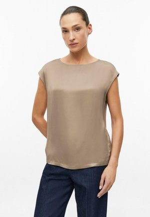 Vrouw met haar in een knot, die een beige blouse met korte mouwen en donkerblauwe broek draagt, staande tegen een witte achtergrond.