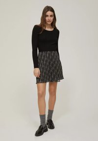 Top lungo a maniche lunghe nero a coste abbinato a una gonna nera con un motivo circolare su tutta la superficie. Il modello indossa calzini grigi e mocassini neri.