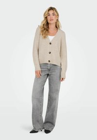 Cárdigan beige de punto con escote en V, cinco botones oscuros y puños acanalados, combinado con jeans gris de pierna ancha y tacones negros de punta.