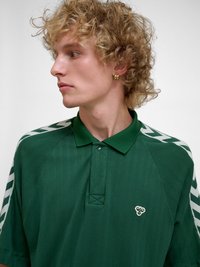 Ung person med krøllet blondt hår iført en mørkegrøn polo-shirt med hvide zigzag-mønstre på ærmerne og et lille logo på brystet.