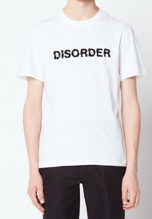 Osoba w białym t-shircie z krótkim rękawem, na którym widnieje czarny, ząbkowany tekst "DISORDER" na piersi, w połączeniu z czarnymi spodniami.
