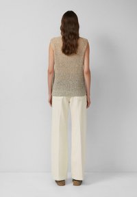 Frau steht mit dem Rücken zugewandt und trägt ein ärmelloses beigefarbenes Strickoberteil, off-white weit geschnittene Hose und braune Schuhe vor einem schlichten Hintergrund.