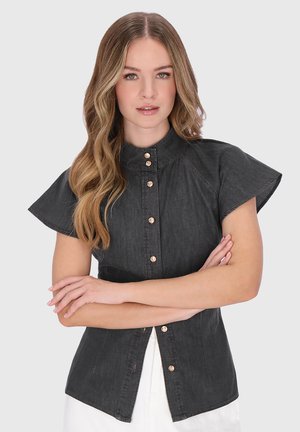 Młoda kobieta z długimi, falującymi blond włosami, ubrana w czarną, krótkorękawową koszulę na guziki, ze skrzyżowanymi ramionami, stojąca na tle jasnoszarego tła.