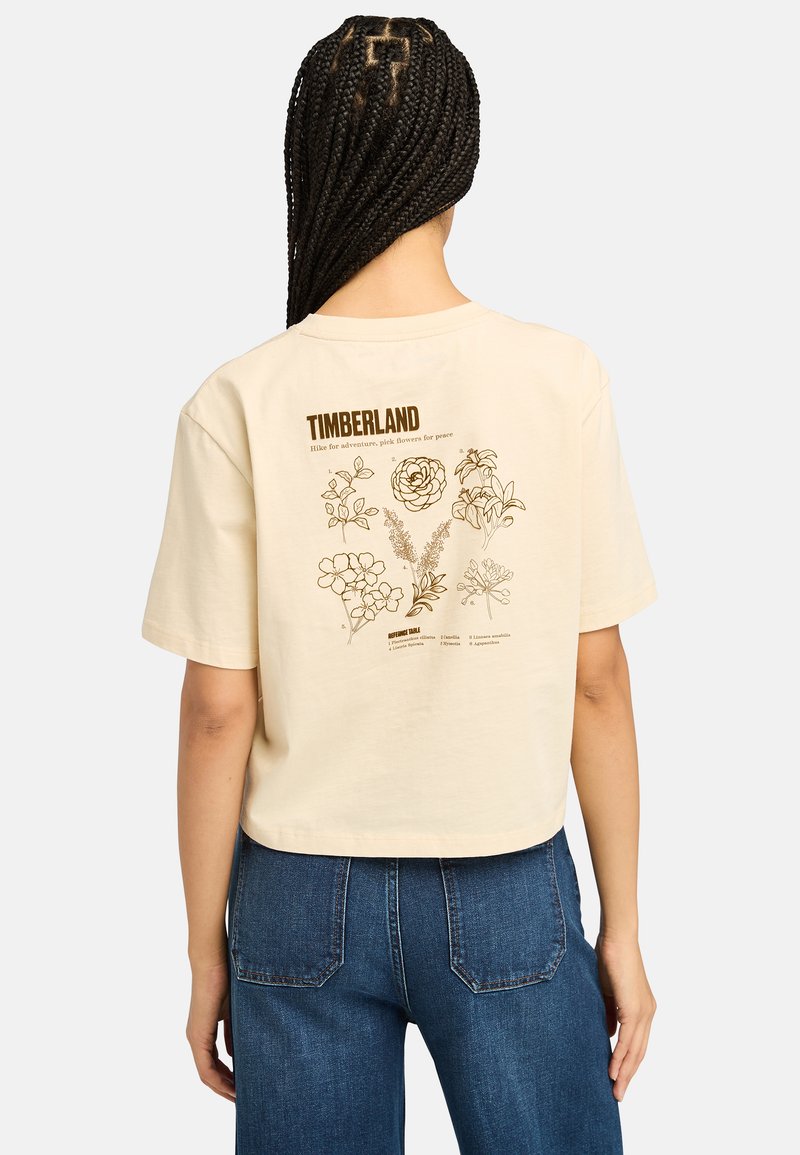 Béžové zkrácené tričko s hnědými květinovými grafikami a nápisem "TIMBERLAND" na zadní straně. Krátké rukávy, měkký bavlněný materiál.