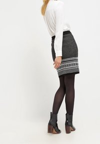 Jupe noire avec un motif géométrique blanc à l'ourlet, avec une coupe ajustée. Portée avec des collants noirs et des bottines à talons. Blouse blanche visible.