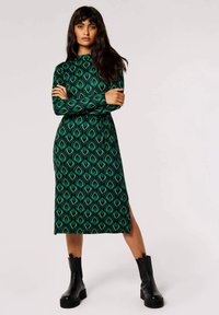Abito verde a maniche lunghe con motivo a foglia, spacco laterale e scollatura alta. Indossato con stivali neri alla caviglia, caratterizzati da una silhouette aderente.