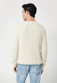 Maglione lavorato a maglia color crema con motivo a trecce, caratterizzato da un collo rotondo e polsini a coste, indossato sopra jeans blu chiaro.