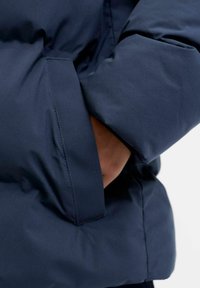 Tumši zila pūkaina jaka ar maigu, gludu tekstūru. Ir sānu kabata un pūkaina forma, kas raksturīga aukstam laikam piemērotai apģērba veidam.