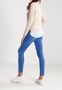 Pull beige texturé, chemise blanche superposée en dessous, jean skinny bleu et baskets blanches. Accent sur la coupe ajustée et le tissu lisse.