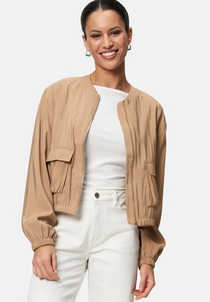 Beige lightweight bomber jacket with a zip front, elastic cuffs, and two patch pockets; paired with a white top and white pants.