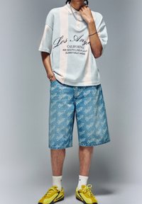 Světle modrý pruhovaný polokošile s bílými detaily a logem, doplněná krátkými džínovými šortkami s opakujícím se vzorem loga. Žluté sportovní boty.