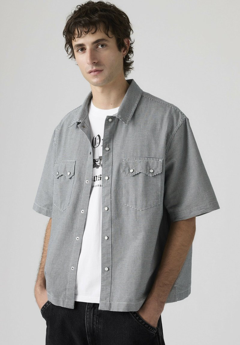 Giovane uomo con capelli ricci e scuri che indossa una camicia a maniche corte a quadri bianca e nera sopra una t-shirt bianca con grafica, con le mani in tasca.