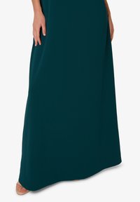 Tyrkysové maxi šaty vyrobené z hladké tkaniny, představují volný siluetu, se rovnými lemy a minimem detailů. Sandály jsou viditelné dole.