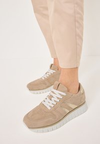 Beige mokkasiner av semsket skinn med hvite lisser, teksturerte detaljer og en tykk hvit såle. Har perforeringer og metalliske striper på sidene.