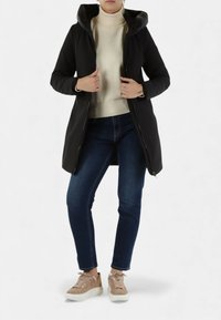 Cappotto nero con cappuccio dalla finitura liscia, chiusura con zip e tasche laterali, indossato sopra un maglione crema e jeans blu, completato da sneakers beige.