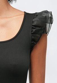 Bezramkowy czarny top z przezroczystymi falbaniastymi rękawkami na osobie o średnim odcieniu skóry na jednolitym tle.