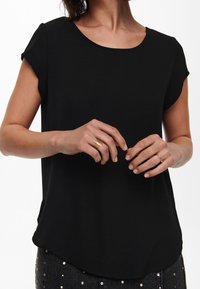 Top negro de manga corta con cuello redondo y una textura suave. El dobladillo es ligeramente curvado y se combina con jeans oscuros adornados.