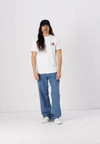 Mann med langt hår og bart iført svart caps, hvit T-skjorte med logo, løse blå jeans og hvite sneakers stående mot hvit bakgrunn.