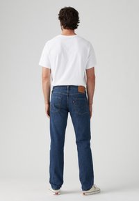 Jeans de mezclilla azul con corte recto, que cuentan con bolsillos traseros y un parche de cuero marrón en la cintura, combinados con una camiseta blanca.