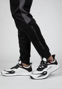 Zwarte sportieve broek met grijze zijpanelen, voorzien van de tekst "GORILLA WEAR". Zwart-witte sportieve schoenen met rode accenten en een gestructureerd materiaal.