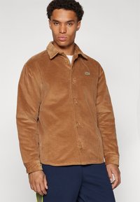 Brown corduroy shirt with a button-up front, pointed collar, and a small green logo on the left chest. Soft texture, ribbed design.