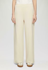 Pantalons larges, beige clair, avec une texture lisse et une taille élastique, associés à des chaussures de couleur claire. Design simple et minimaliste.