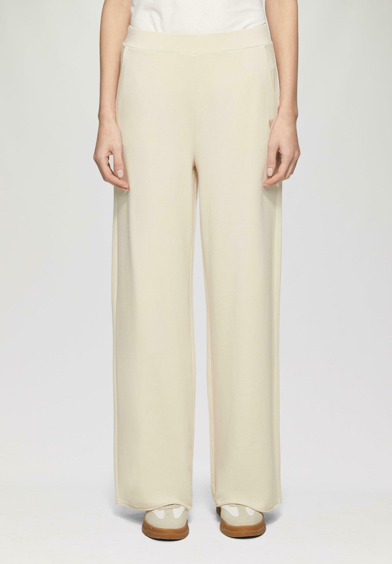 Pantalons larges, beige clair, avec une texture lisse et une taille élastique, associés à des chaussures de couleur claire. Design simple et minimaliste.