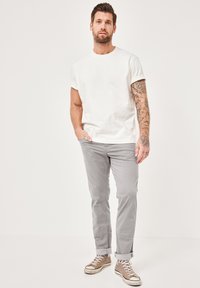 White short-sleeve t-shirt, light gray pants, brown sneakers with rolled cuffs. Man stands with one hand in pocket, minimal background.