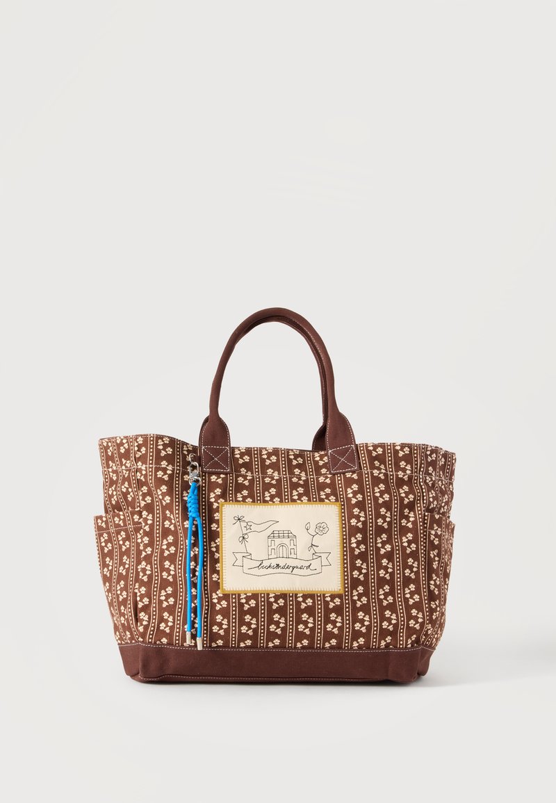 Sac cabas marron à motif floral avec deux anses, poches latérales et un écusson avant représentant une maison et un texte décoratif.