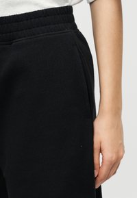 Κοντινό πλάνο μέσης και χεριού ατόμου που φοράει μαύρο λαστιχένιο παντελόνι φόρμας με πλαϊνές τσέπες και λευκό πουκάμισο.