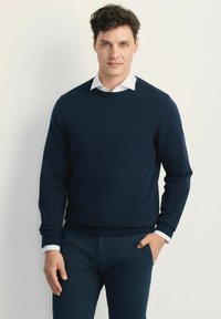 Granatowy sweter z dzianiny z okrągłym dekoltem, noszony na białej koszuli z kołnierzykiem. Ręce włożone w kieszenie.