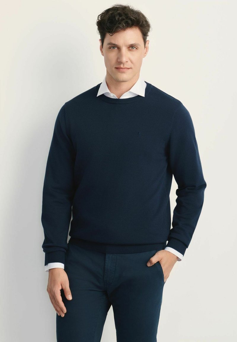 Granatowy sweter z dzianiny z okrągłym dekoltem, noszony na białej koszuli z kołnierzykiem. Ręce włożone w kieszenie.