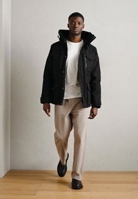 Chaqueta negra aislante con capucha, cierre frontal y múltiples bolsillos; combinada con una camiseta blanca y pantalones beige claros. Zapatos negros.