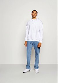 Sudadera con capucha blanca y bolsillo delantero, jeans de color azul claro y zapatillas blancas. El modelo está de pie contra un fondo neutro.